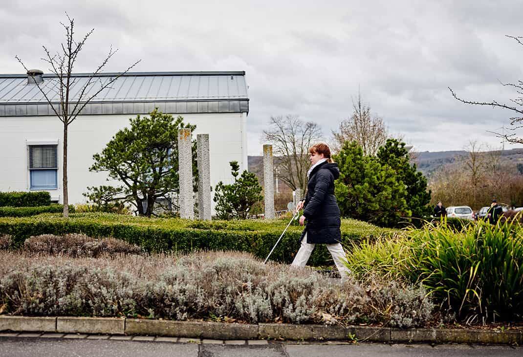 A woman (Sandra Pilz) who is blind using her white cane to walk on the green grounds of the BFW campus, with rolling hills in the background.